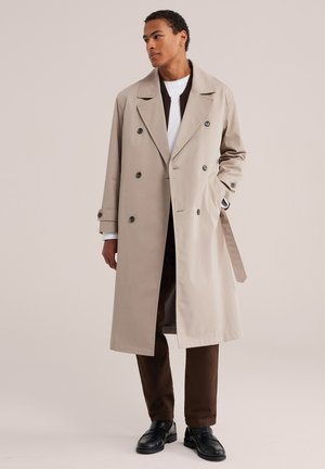Jeune homme portant un trench-coat beige à double boutonnage, une chemise blanche, un pantalon marron et des chaussures noires, debout avec une main dans la poche.