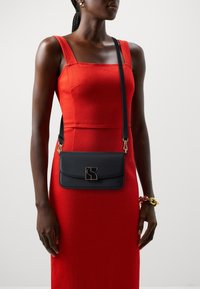 Black crossbody bag made of leather with a gold geometric logo. The bag features a flat design and an adjustable strap.