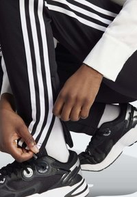 Černé sportovní kalhoty s bílými pruhy, vybavené zipem na lemu. Černé sportovní boty s metalickými stříbrnými akcenty a bílou podrážkou. Viditelné bílé ponožky.