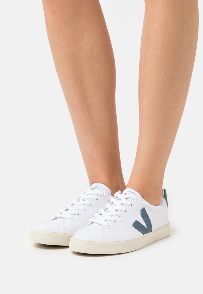 Sapatilhas brancas de lona com um acabamento texturizado, apresentando detalhes cinzentos e uma sola de borracha. Os atacadores e um logotipo na lateral completam o design.