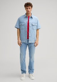 Camicia di jeans azzurro chiaro a maniche corte con due tasche sul petto, indossata sopra una maglietta rossa e abbinata a jeans azzurro chiaro e sneakers bianche.