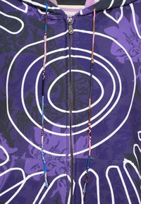 Felpa con zip viola con motivi a spirale bianchi, dotata di coulisse texturizzate in vari colori. Il tessuto è morbido e con un design grafico.