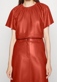 Haut en cuir rouge à manches courtes avec un froncé au niveau du col, assorti à une jupe taille haute en cuir avec une ceinture visible.