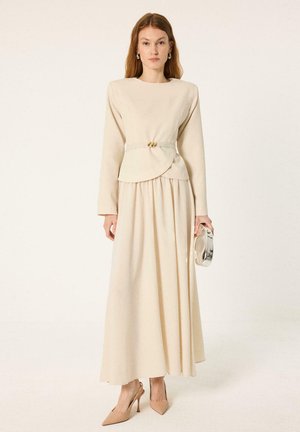Donna che indossa un lungo abito beige con spalle strutturate e dettaglio cintura, tiene una pochette argentata e indossa décolleté nude con punta.