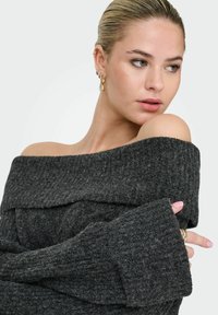 Sweter w kolorze ciemnoszarym, odsłaniający ramiona, o prążkowanej strukturze, z szerokimi rękawami i fałdowanymi detalami przy dekolcie. Widoczne złote i srebrne kolczyki.