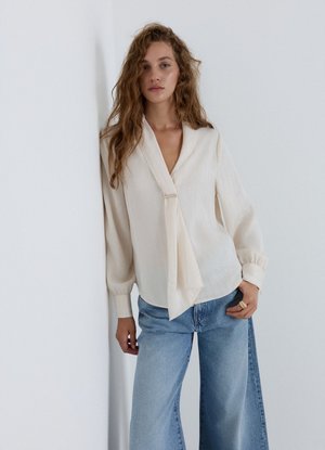 Junge Frau mit langen, welligen Haaren, die eine cremefarbene Bluse mit Bindedetail und weit geschnittene blaue Jeans trägt, vor einer weißen Wand stehend.
