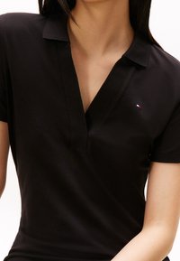 Schwarzes Polo-Shirt mit kurzen Ärmeln, V-Ausschnitt-Design und subtilen Logo-Details in Rot, Weiß und Blau im linken Brustbereich. Strukturierten Stoff.