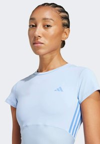 Chemise de sport bleu clair à manches courtes, texture côtelée à la taille, et design signature à trois bandes de couleur assortie sur le côté.