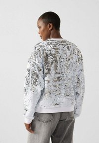 Persoon die een zilver-witte top met pailletten en grijze spijkerbroek draagt, zijwaarts staand tegen een effen lichte achtergrond.