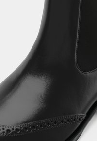 Cizme Chelsea din piele neagră, cu perforații brogue și panou elastic lateral, prezentate în prim-plan pe un fundal alb.