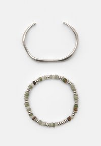 Srebrna bransoletka na mankiet o lekko nieregularnym kształcie, nad elastyczną bransoletką z koralików w kolorach zielonym, białym, brązowym i srebrnym na białym tle.