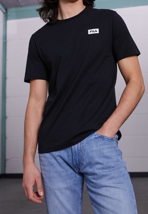 Schwarzes Baumwoll-T-Shirt mit kurzen Ärmeln und einem weißen, rechteckigen Logopatch auf der linken Brust. Kombiniert mit hellblauen Jeans.