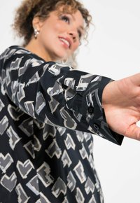 Camicia di tessuto nera con un motivo geometrico in grigio e bianco. Le maniche lunghe presentano dettagli con bottoni e una vestibilità comoda.