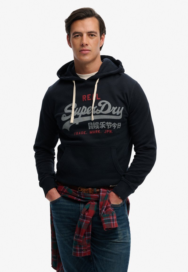 Homme portant un sweat à capuche bleu marine SuperDry avec des cordons blancs, une chemise à carreaux rouge nouée autour de la taille et un jean bleu, debout les mains dans les poches.