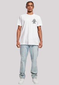 White cotton t-shirt featuring a black graphic design on the left chest. Paired with light blue denim jeans and white sneakers.