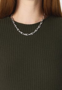 Stříbrný řetízek s oválnými články a jemnou texturovanou úpravou, odpočívající na pruhovaném tmavě zeleném topu.