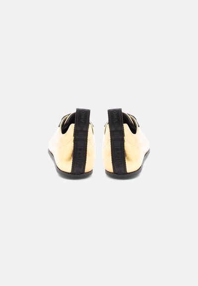 Pár lesklých zlatých topánok zobrazených zozadu, každá s čiernym vertikálnym pútkom a čiernou podrážkou na jednoduchom pozadí.