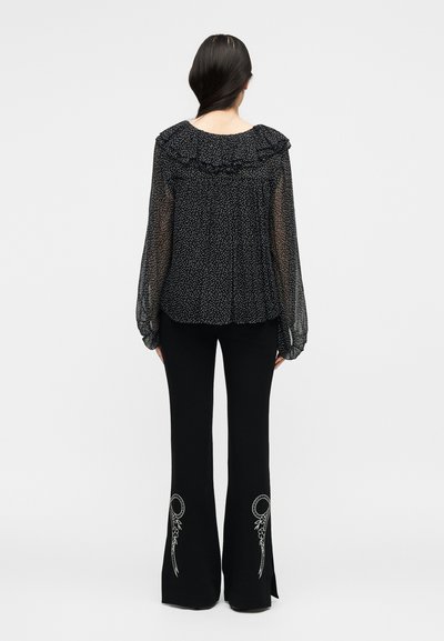 Γυναίκα με μακριά μαλλιά, φορώντας μαύρο διάφανο πουκάμισο με πουά και μαύρο παντελόνι με κεντήματα, φωτογραφημένη από πίσω.