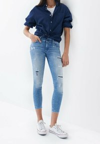 Jeans de mezclilla azul claro con detalles desgastados, corte ajustado, cintura alta y longitud hasta el tobillo. Combinados con una camisa azul marino y zapatillas blancas.