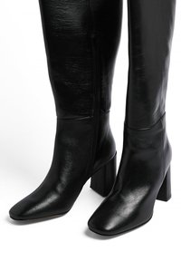 Black knee-high boots made of shiny leather, featuring a square toe and block heel. Side zipper and smooth texture.