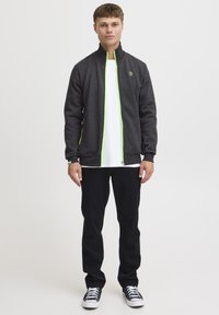 Chaqueta gris con cremallera y acentos verdes, combinada con una camisa blanca y pantalones negros, llevada por una persona de pie contra un fondo liso.