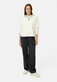 Heller, cremfarbener Fleece-Pullover mit halb verdecktem Reißverschluss, langen Ärmeln, kombiniert mit dunklen, weiten Jeans und hellen Turnschuhen.