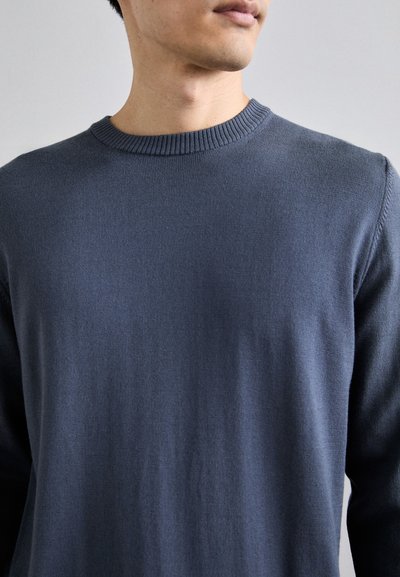 Un pull bleu marine à col rond avec un col côtelé, une texture lisse, des manches longues et une coupe ajustée, faisant apparaître un style léger et minimaliste.