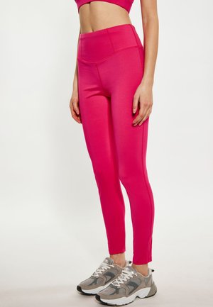 Kvinde iført lyserøde højtaljede leggings og grå og hvide sneakers, stående mod en ensfarvet lys baggrund.