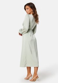 Dlouhé rukávy, mátově zelené šaty s úzkým pasem, hladká textura a květinová výšivka na rukávech. Délka do poloviny lýtek. Průhledné sandály na podpatku.