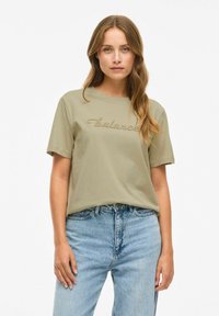 Olijfgroene katoenen t-shirt met korte mouwen, met geborduurde tekst "balance" op de borst. Draag het met lichtblauwe denim jeans.