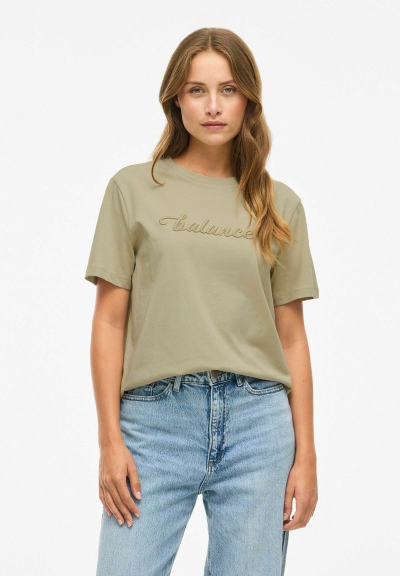 Olijfgroene katoenen t-shirt met korte mouwen, met geborduurde tekst "balance" op de borst. Draag het met lichtblauwe denim jeans.