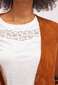 Hnědá semišová bunda přes bílou košili s dekorační krajkovou vzorovou výšivkou na hrudi. Materiály jsou měkké s hladkým povrchem.