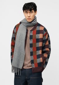 Νεαρός άνδρας φοράει πολύχρωμο καρό ζακέτα, ριγέ πουκάμισο, σκούρο τζιν και παχύ γκρι κασκόλ μπροστά σε απλό φόντο.