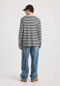 Gestreepte lange mouwen shirt in marineblauw en crème; relaxte pasvorm. Gecombineerd met lichtblauwe denim broek en beige schoeisel; accent van een zwarte pet.