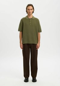 Jeune homme debout portant un polo en maille à manches courtes vert olive, un pantalon large marron foncé et des mocassins noirs sur fond blanc.