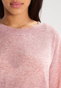 Nära bild av en person som bär en mjuk rosa stickad tröja med rund halsringning, som visar läppar, nacke och övre bröst mot en ljus bakgrund.