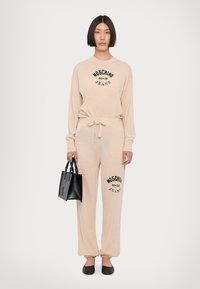 Conjunto de roupa de casa bege com o logótipo preto "Moschino Jeans". Sweater de mangas longas, calças de moletom com cordão, acompanhadas por uma pequena bolsa preta.