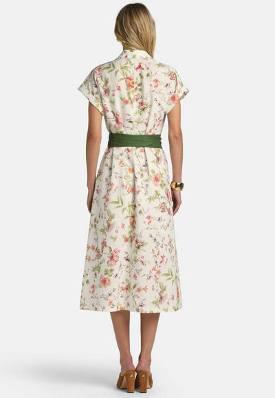 Mujer con vestido midi blanco con flores, mangas cortas, cinturón verde en la cintura, pulsera dorada y zapatos marrones de tacón, de espaldas.