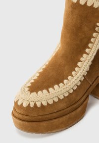 Stivale alla caviglia in suede marrone con dettagli intrecciati color crema lungo la cucitura e una suola spessa e imbottita.