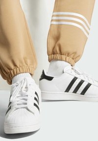 Beige joggers with elastic cuffs and three white stripes on the side paired with white sneakers featuring black stripes and a textured toe cap.