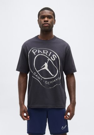 Férfi fekete Paris Saint-Germain pólóban és sötétkék Nike rövidnadrágban, egy sima világos háttér előtt áll.