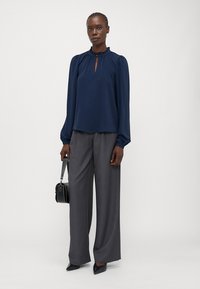 Blusa blu navy con scollatura tonda, dettagli arricciati e apertura a chiave, abbinata a pantaloni grigi a gamba larga e décolleté neri a punta.