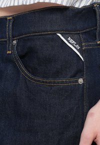 Temno modre hlače iz jeansa z rumenim kontrastnim šivom, majhnim sprednjim žepom in etiketo z napisom "REPLAY." Opremljene s kovinskimi dodatki.