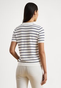 Svetlokrémový krátky rukáv s námorníckymi pruhmi, s rebrovaným golierom a lemom. Modelka má na sebe priliehavé svetlé nohavice.