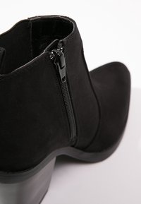 Bottine en daim noir avec un bout pointu, une fermeture à glissière latérale et un talon chunky, présentant une texture lisse et un design minimaliste.