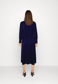 Vestido azul marinho de mangas longas com cintura ajustada, saia fluida e detalhes de botões nos punhos. Textura suave, silhueta até ao joelho.