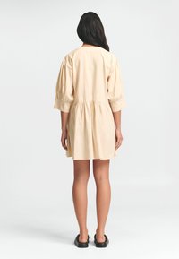 Kurzärmliges, hellbeiges Kleid aus Baumwolle mit eingeschnürter Taille, knielangem Rock und weichem Material. Besitzt einen runden Ausschnitt.