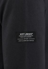 Tela negra con una superficie texturizada que presenta una etiqueta rectangular. La etiqueta tiene texto en blanco, indicando la marca y su año de fundación.