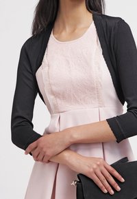 Svart cardigan över en ljusrosa klänning med en texturerad spetsliv och veckad kjol. En svart handväska hålls i händerna.