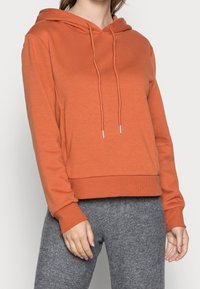 Oranžová bavlněná mikina s kapucí, přední klokaní kapsou, stahovací šňůrkou a žebrovanými manžetami a lemem. V kombinaci se šedými tepláky.